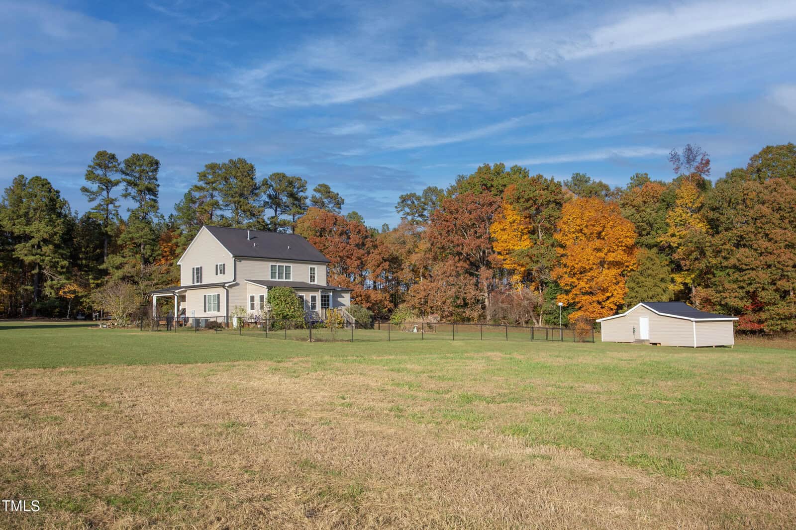 A two-story house surrounded by a grassy yard and colorful autumn trees under a blue sky. A small white shed sits to the right of the house, which is bordered by a black fence.