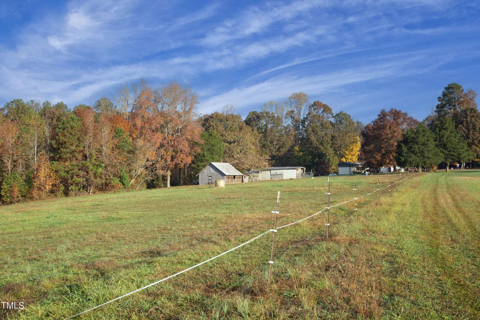 A scenic rural landscape with a grassy field, a few small sheds, and a fence. The background features a line of trees with autumn foliage under a blue sky with wispy clouds.