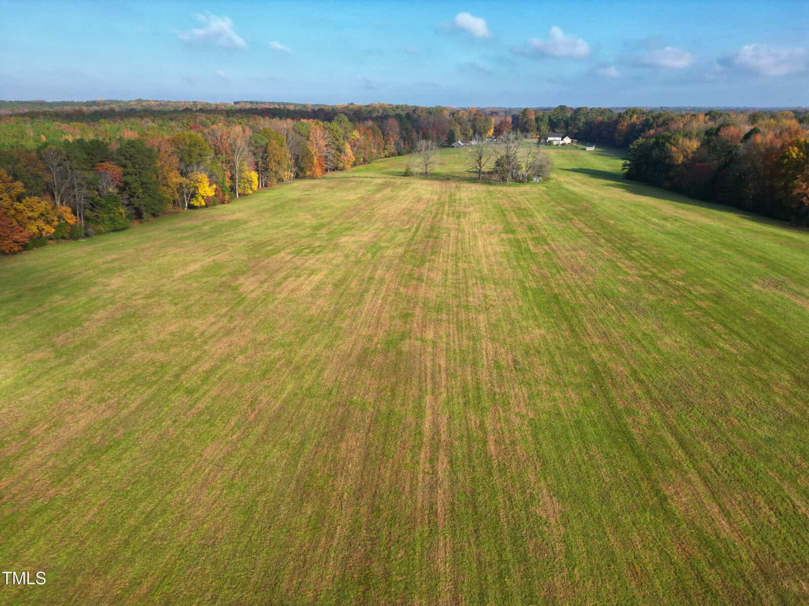 Aerial view of a large, open green field surrounded by trees showing autumn colors. The sky is clear with a few fluffy clouds. In the distance, a few structures are visible at the edge of the field.