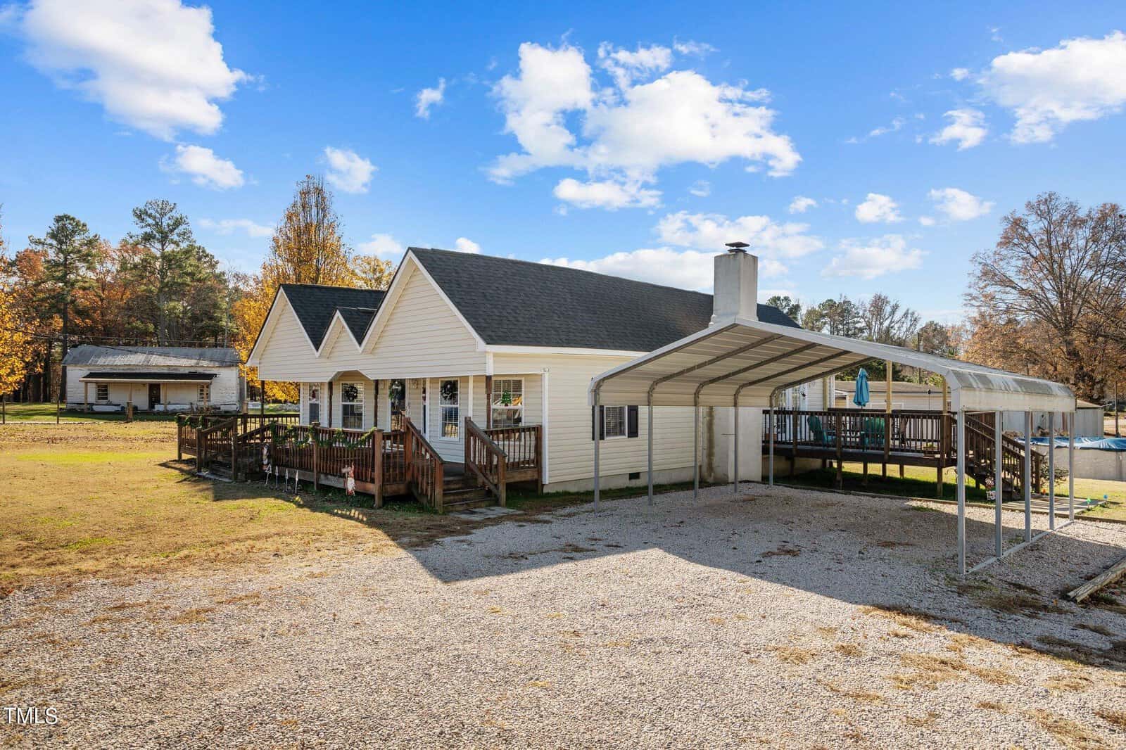 A single-story white house with a gabled roof, featuring a wooden deck and a carport, surrounded by an open yard with sparse trees under a mostly clear sky.