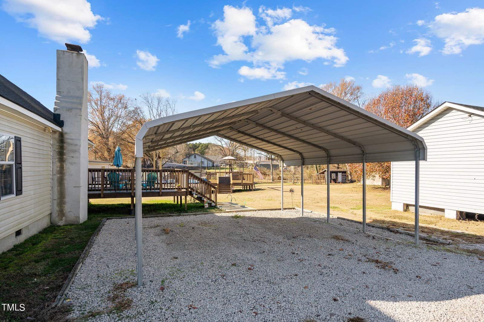 A metal carport installed on a gravel surface beside a suburban home with a wooden deck and a fenced backyard visible in daylight.