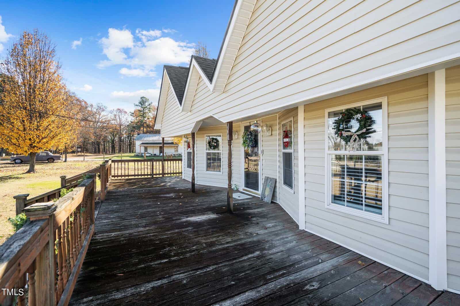 A wooden deck attached to a house with white siding, featuring multiple windows and a glass door with reflections of trees, under a clear sky.