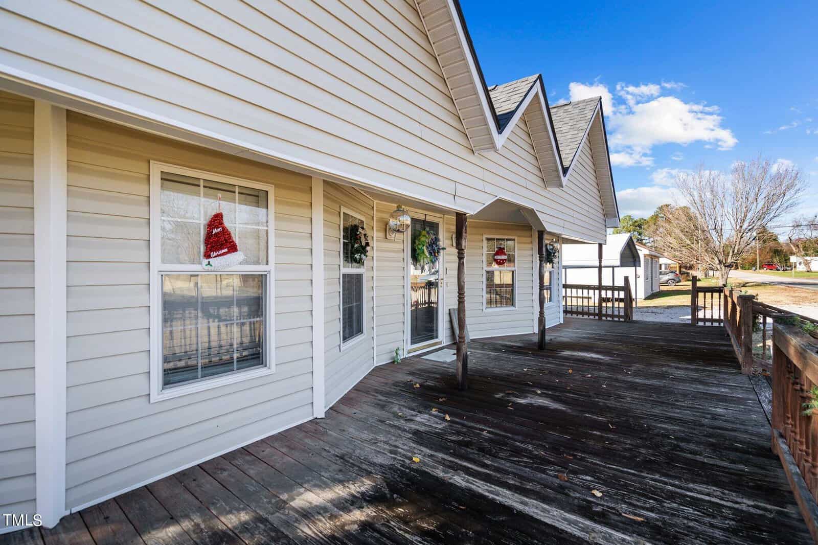 A wooden deck attached to a white-sided house with several windows, some adorned with holiday wreaths, under a clear blue sky.