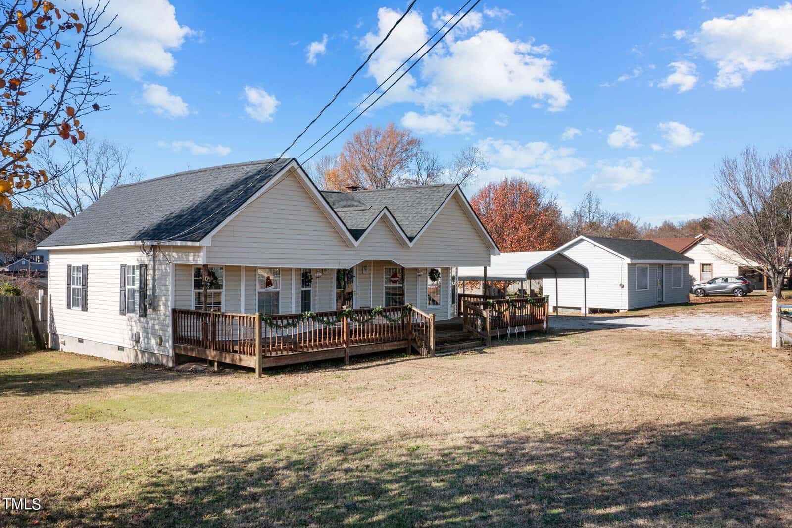 A single-story residential home with white siding features a front deck with wooden railings. a detached structure is visible in the background, and overhead power lines run across a clear sky. the property includes a lawn area in the foreground, devoid of significant vegetation.