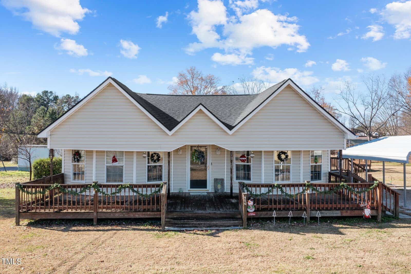 A single-story suburban house with beige siding and a symmetrical gabled roof, featuring a front porch with a wooden railing and festive wreath decorations, situated on a clear day with a well-maintained lawn.