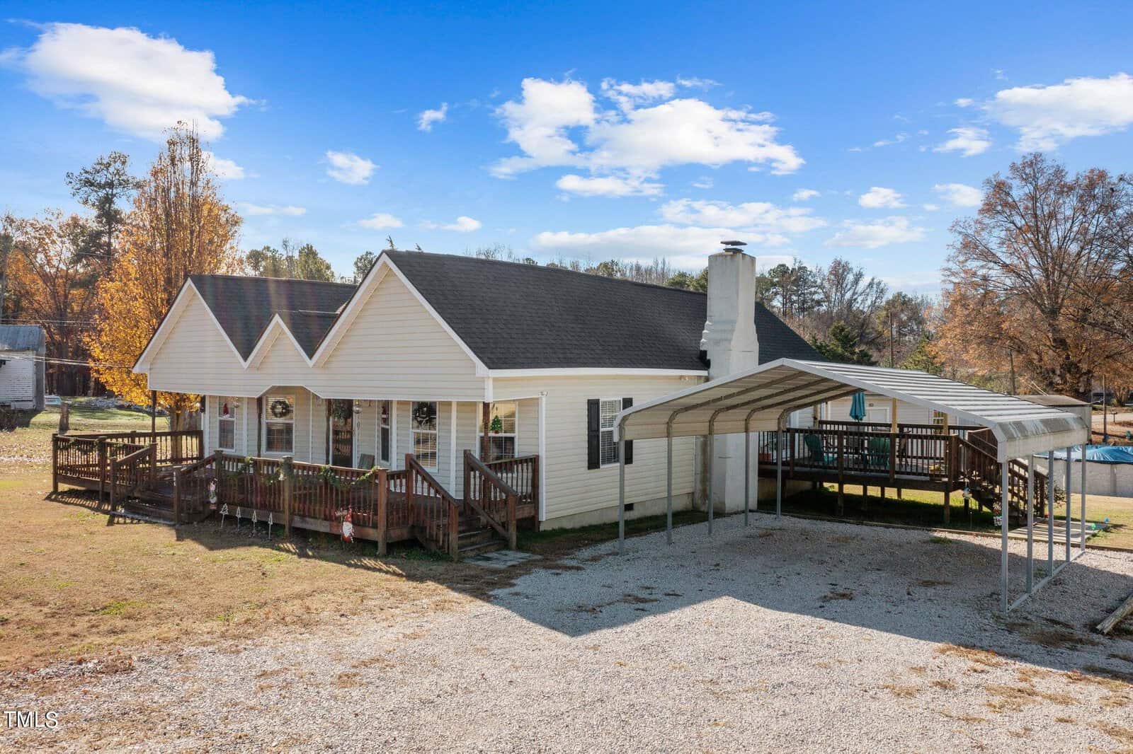 A single-story white house with a steeply pitched roof, featuring an attached carport and a wrap-around wooden deck, set against a backdrop of bare trees and a clear sky.