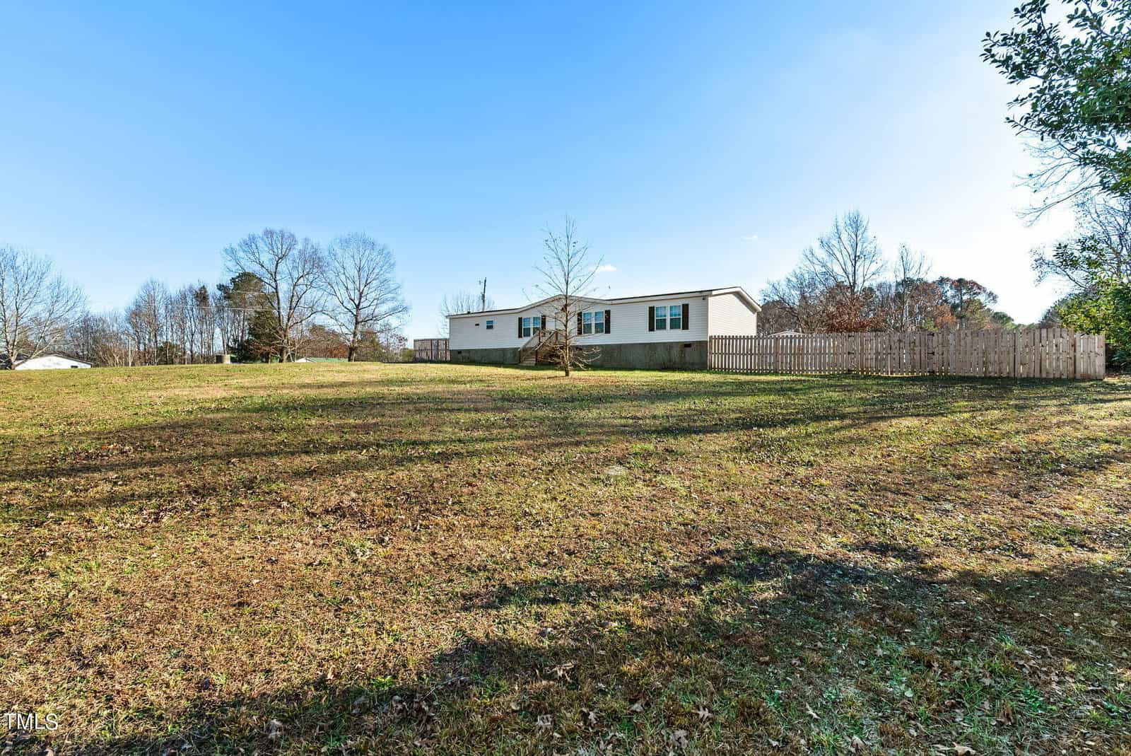 A manufactured home situated on a spacious grassy lot with a wooden fence on one side under a clear blue sky.