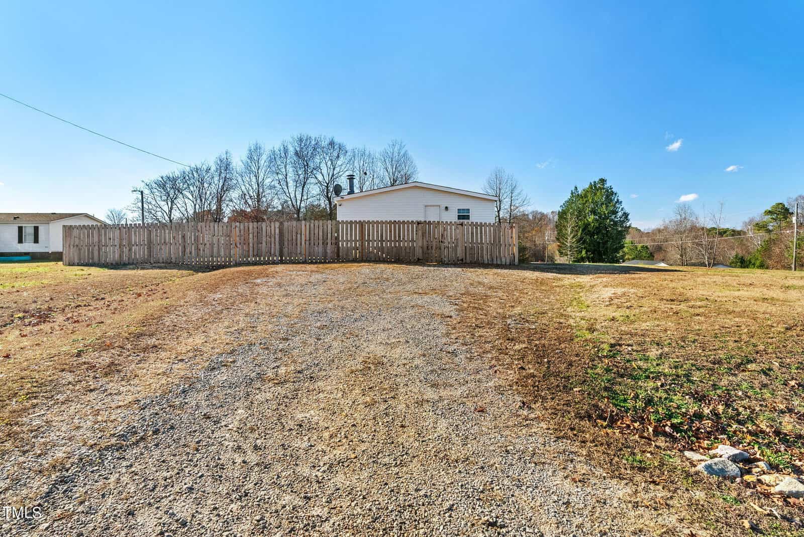A gravel driveway leading to a cream-colored single-story house with a white fenced backyard under a clear blue sky.