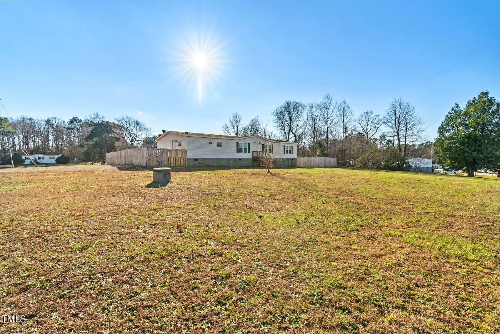A single-story house with a fenced yard under a bright sun in a spacious, grassy field with surrounding trees.