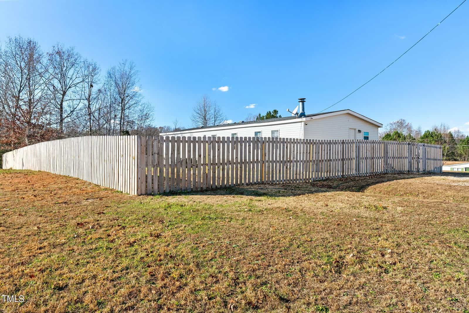 A white mobile home with a pitched roof is enclosed by a tall wooden fence on a sunny day with a clear blue sky.