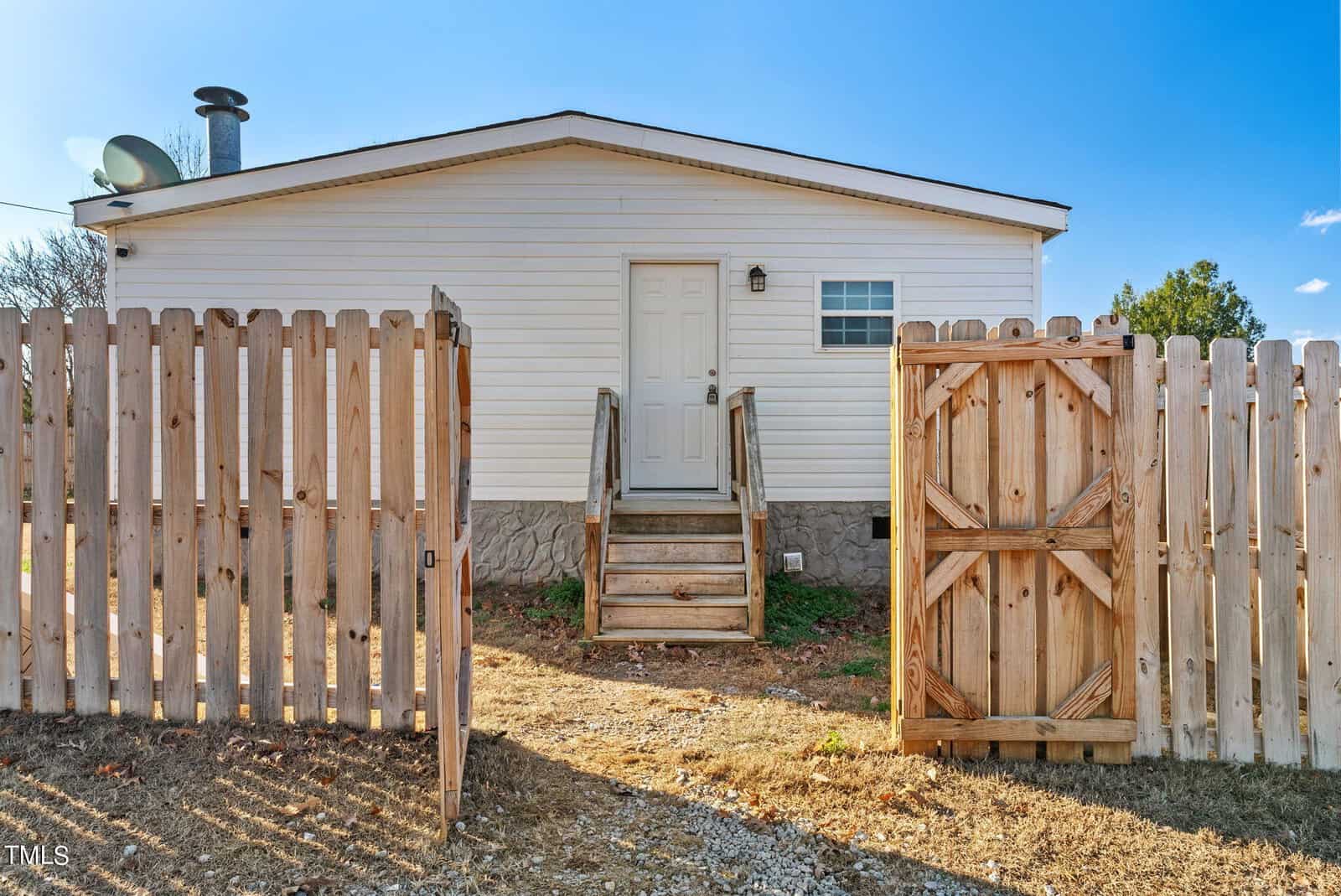 A small white house with a wooden fence gate and gravel driveway under a clear sky.