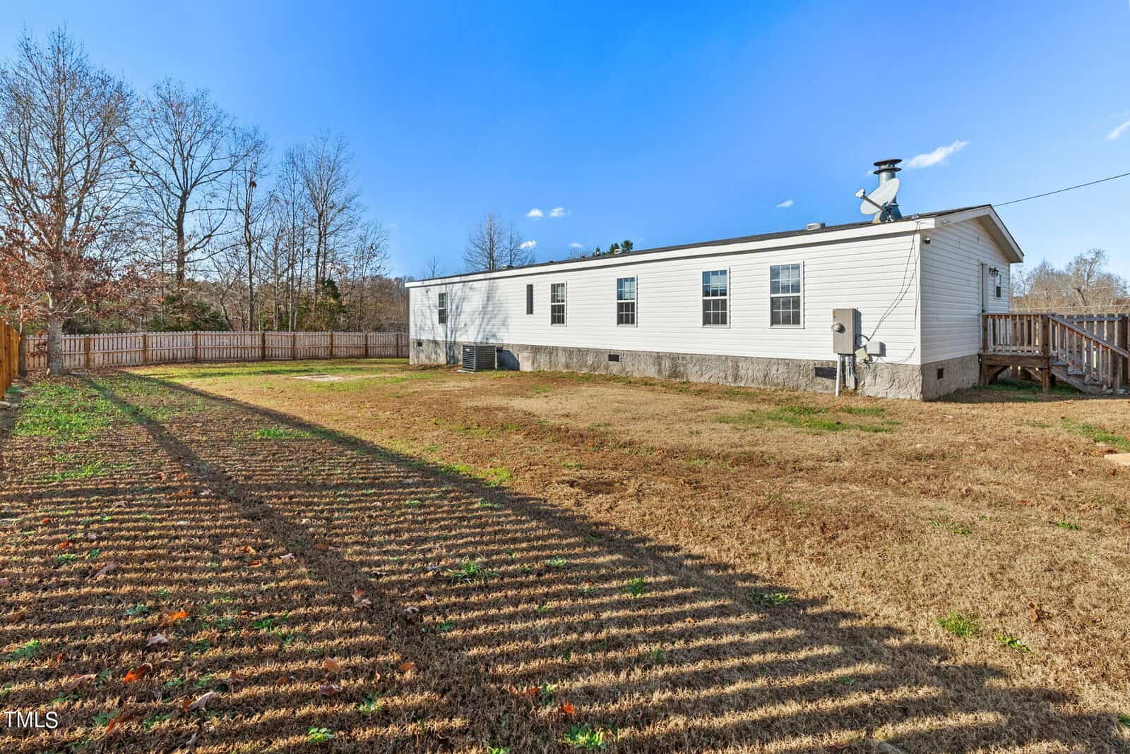 A single-story white mobile home with a wooden deck and a satellite dish on a clear day, enclosed by a wooden fence with a large open yard showing shadow patterns of bare trees.