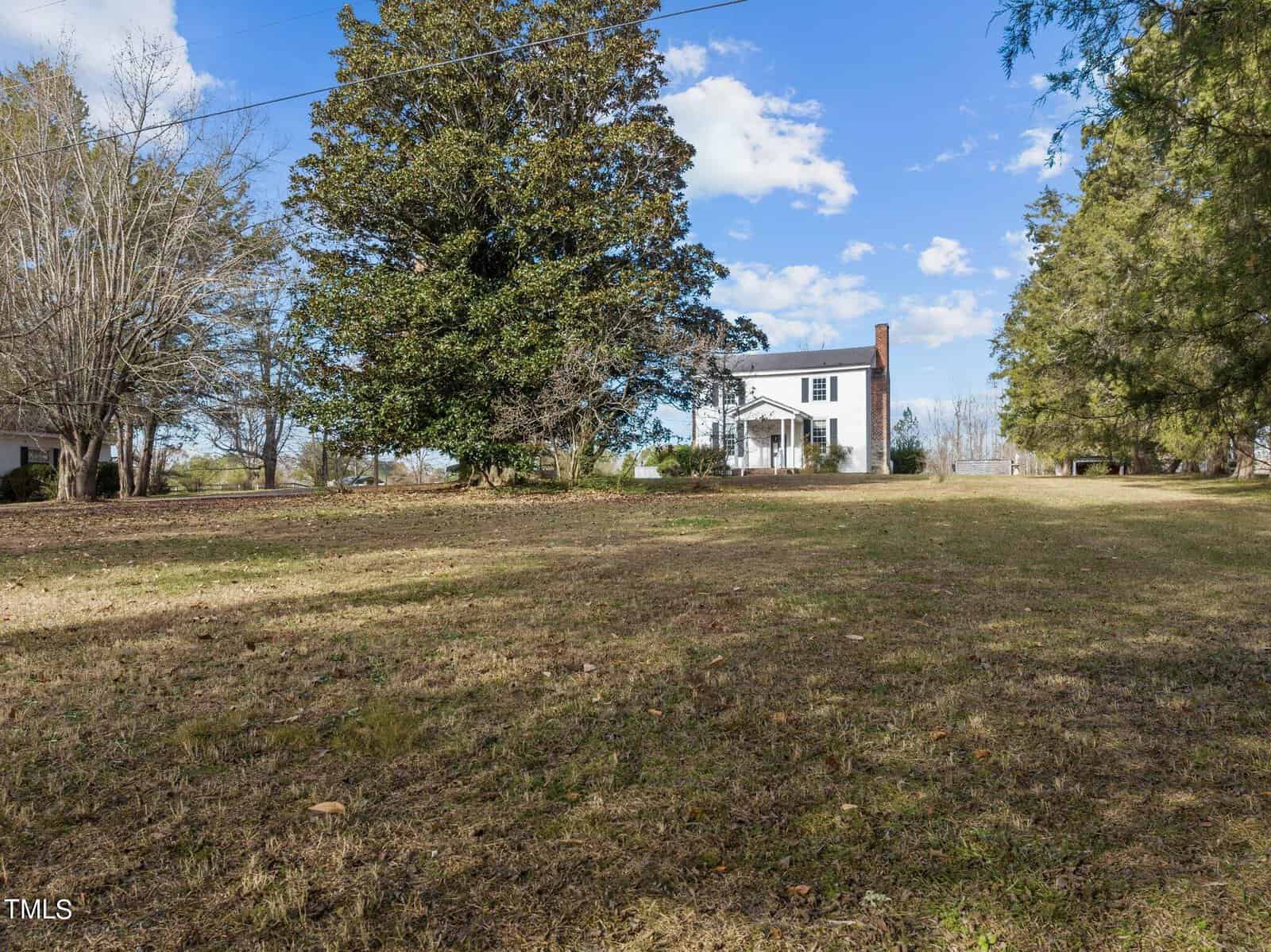 A white two-story house with a chimney is surrounded by trees and a large grassy field under a blue sky with scattered clouds.