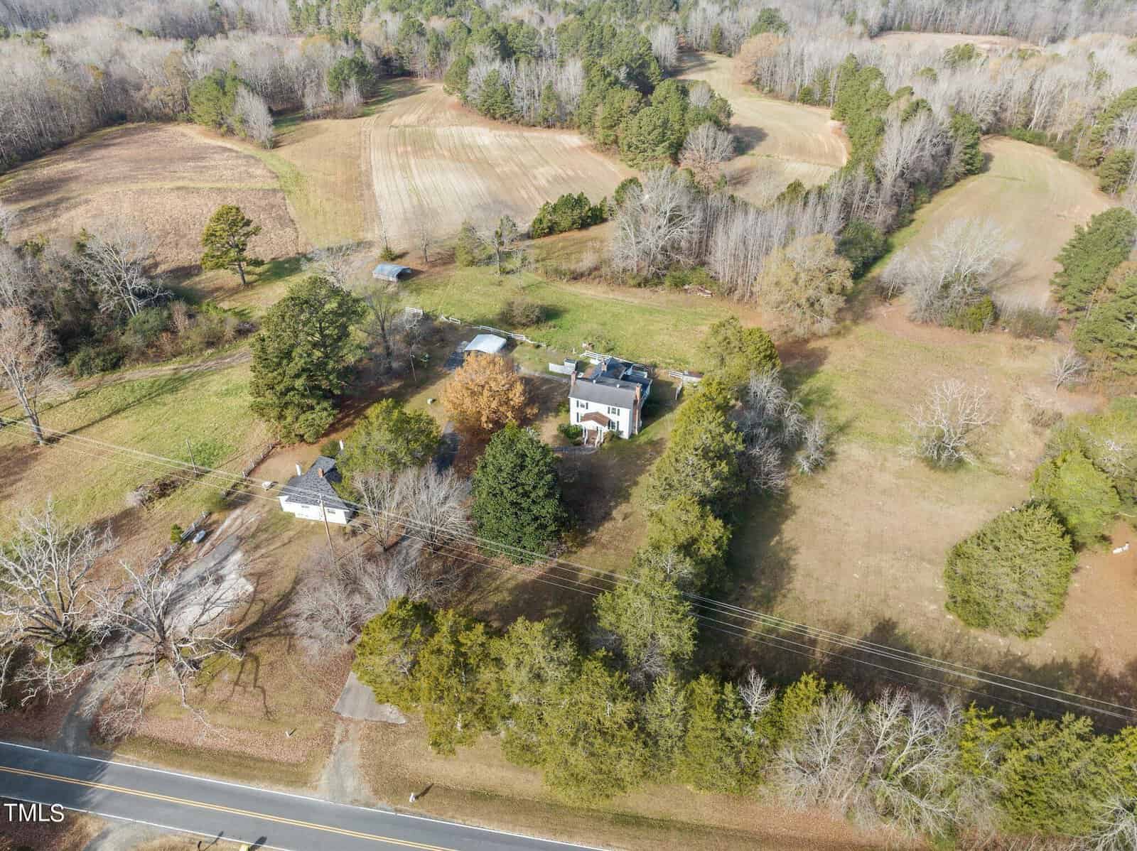 Aerial view of a rural landscape featuring a two-story house surrounded by trees. Nearby are fields, additional small buildings, and a road in the foreground. The area is bordered by wooded patches and open grasslands.