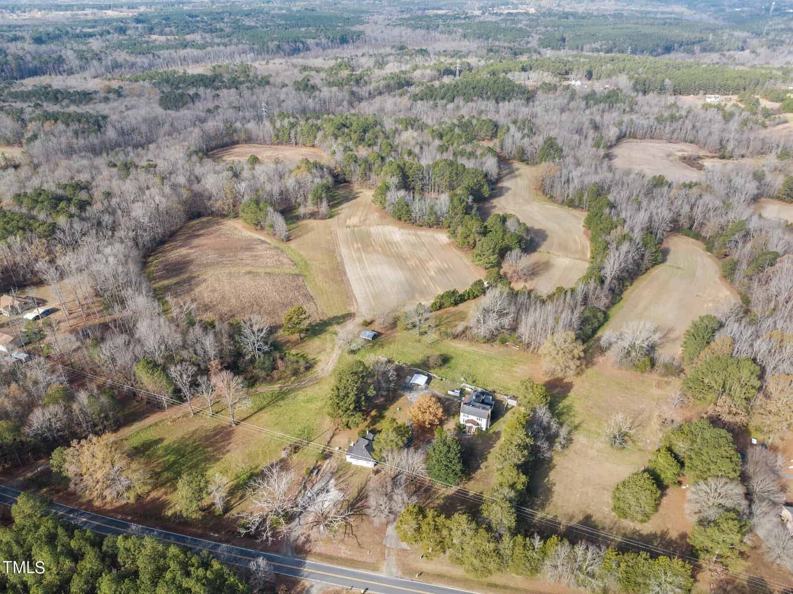 Aerial view of a rural landscape featuring large open fields with patches of trees. A few houses are visible, surrounded by greenery. A road runs horizontally along the bottom of the image, bordered by trees on either side.