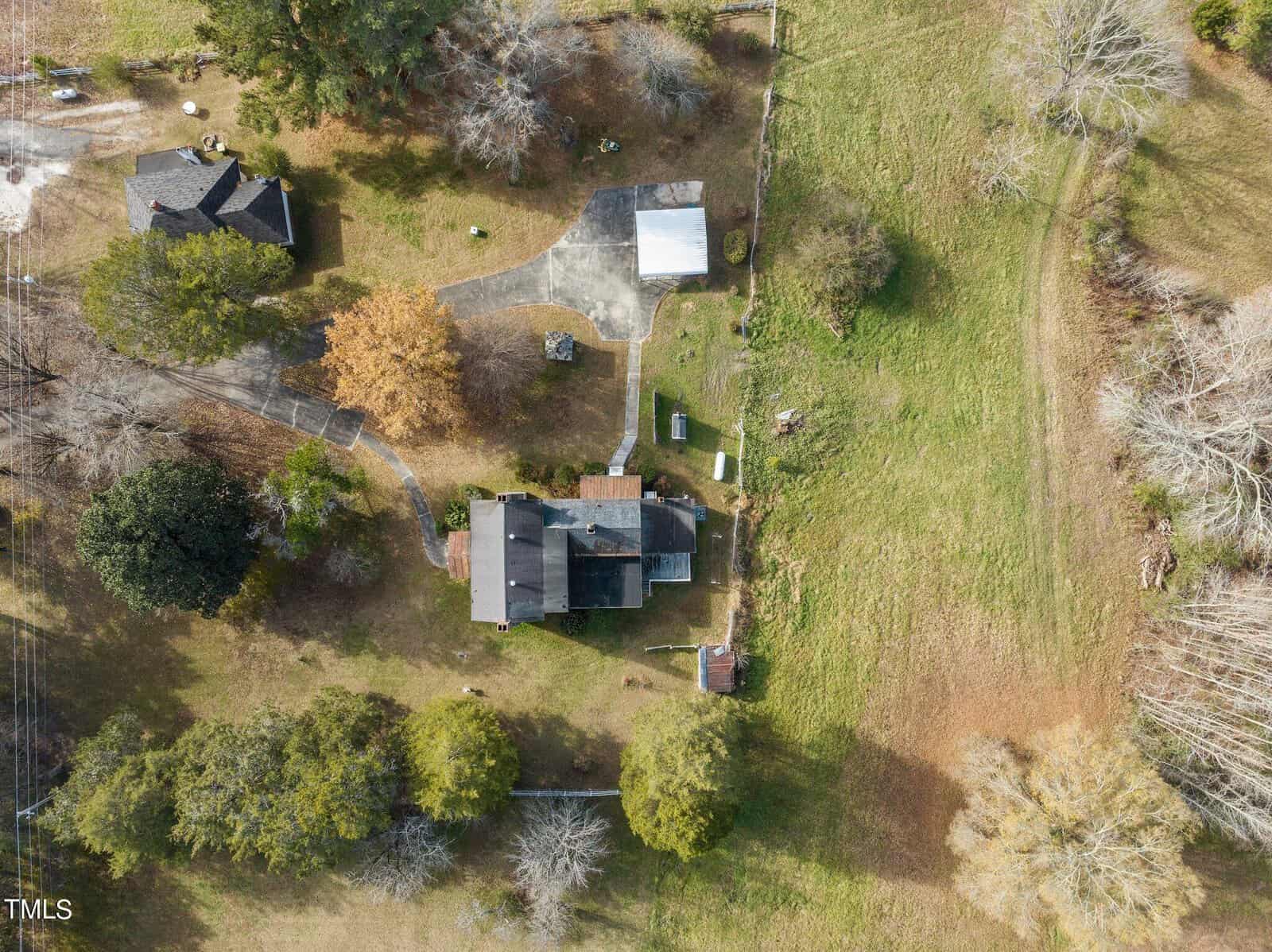 Aerial view of a farmhouse surrounded by greenery. There's a main building with a driveway, several trees scattered around, and neighboring structures. The landscape includes open fields and a small pathway leading from the house.