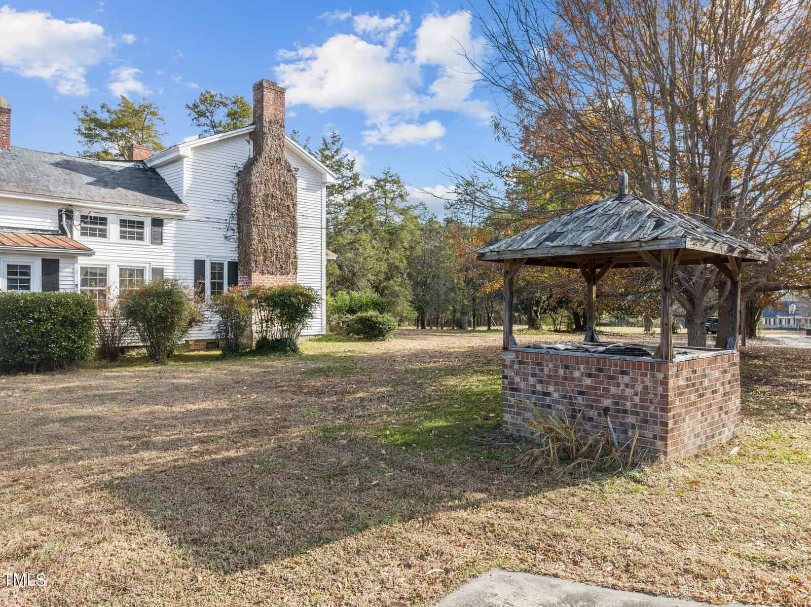 A white house with a large chimney has a spacious yard. In the foreground, there's a brick well structure with a wooden roof. Trees with autumn foliage surround the area, and the sky is partly cloudy.
