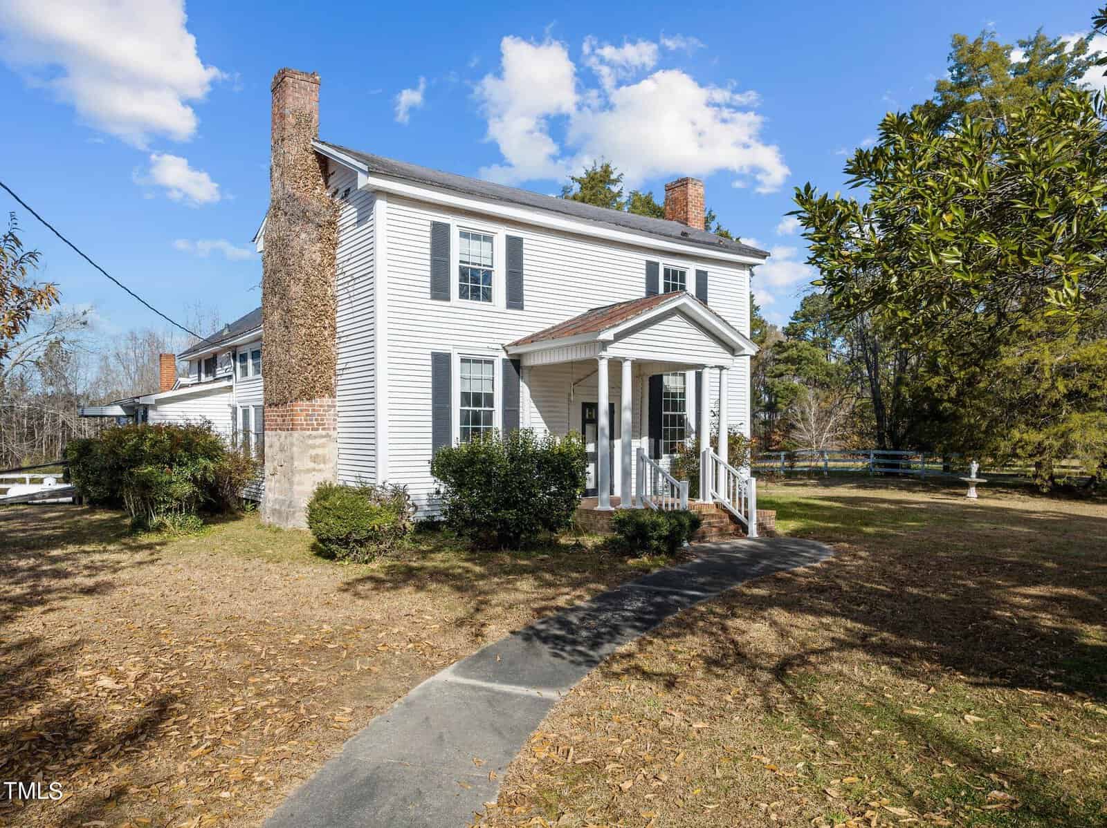 A two-story white house with brick chimneys, a covered porch, and a walkway leading to the entrance. It's surrounded by trees and a lawn under a blue sky with clouds.