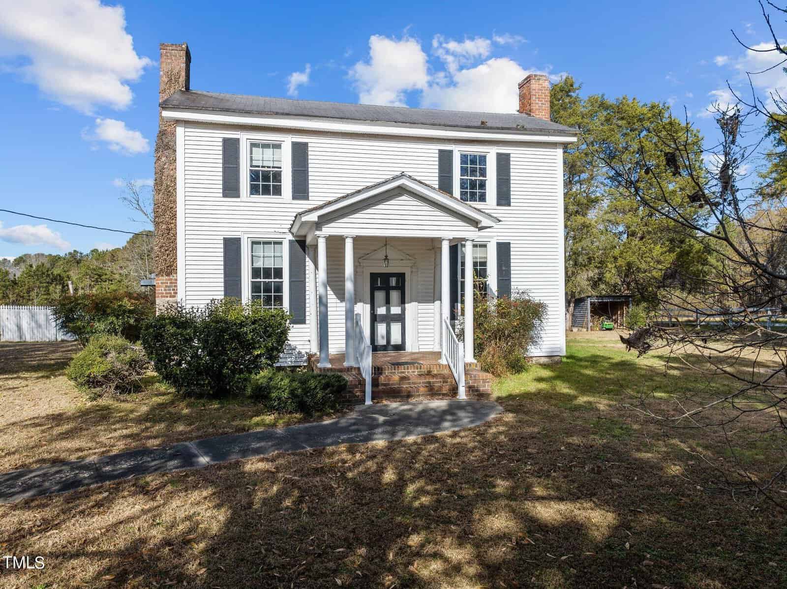 Located at 1579 Pope Road in Creedmoor, this charming two-story white house features a welcoming front porch with columns, nestled amid trees and shrubs. A picturesque pathway leads to the front door under a clear sky dotted with a few clouds.