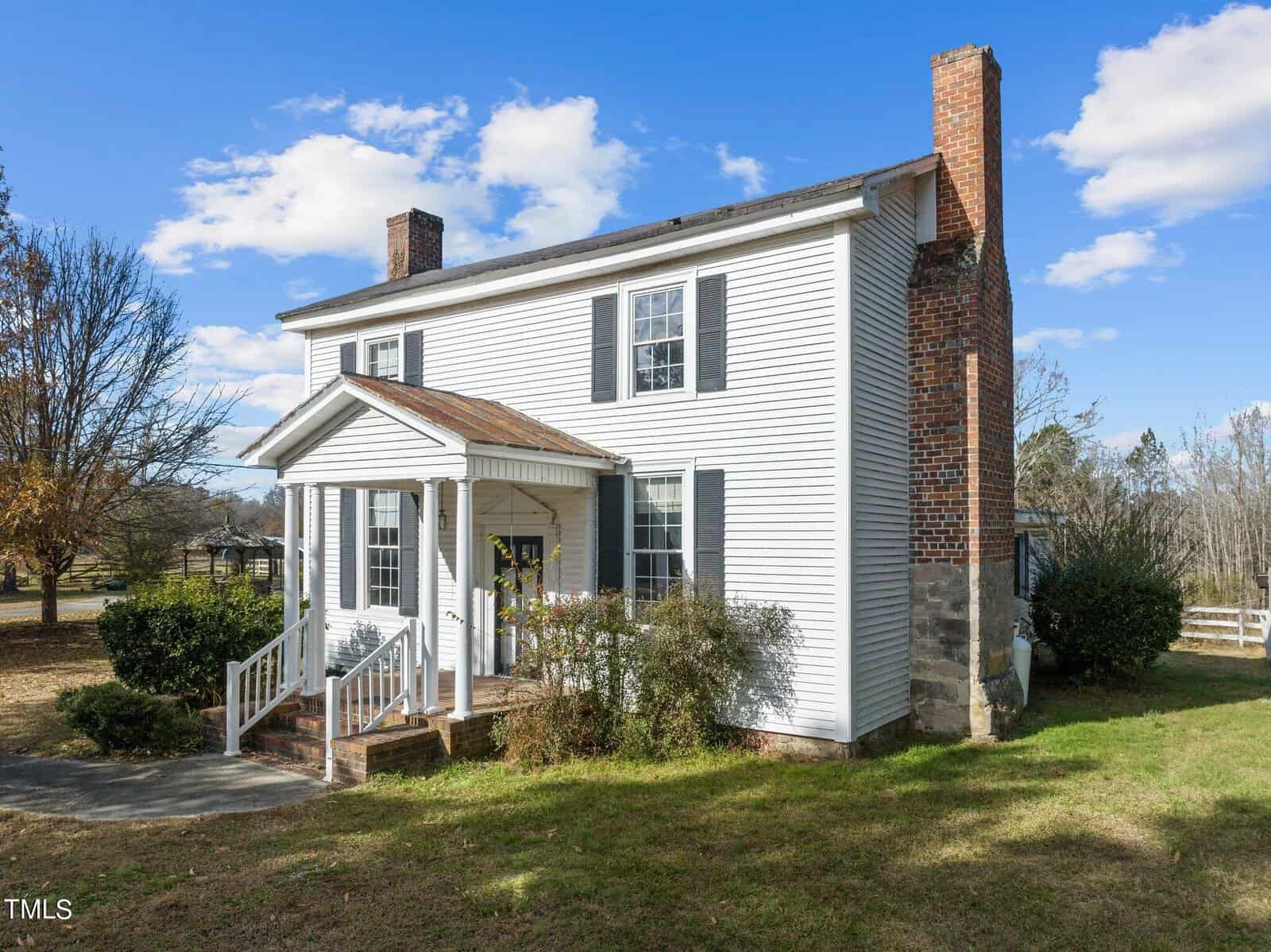 A white, two-story house with a brick chimney and front porch, surrounded by trees and grass under a blue sky with scattered clouds. A pathway leads to the entrance flanked by shrubs.