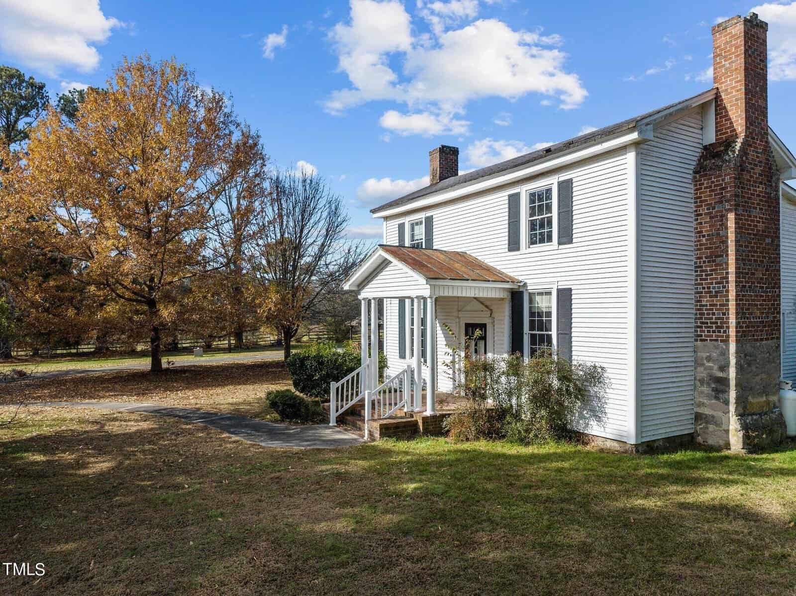 A two-story white wooden house with a brick chimney and a small front porch, surrounded by trees with autumn foliage. The sky is clear with a few clouds, and the yard is grass-covered.
