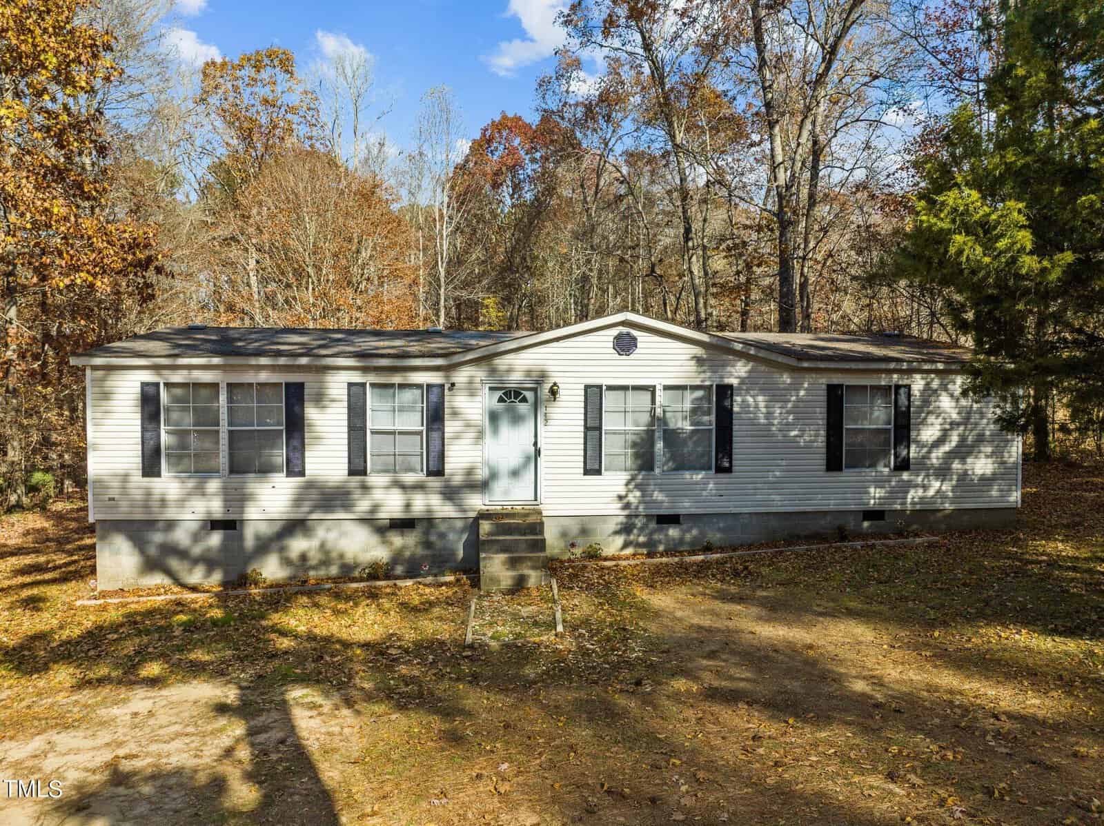 A single-story suburban manufactured home at 142 Village Drive, Henderson NC, with a white exterior, featuring multiple windows and a central front door, set against a backdrop of a wooded area with fallen leaves