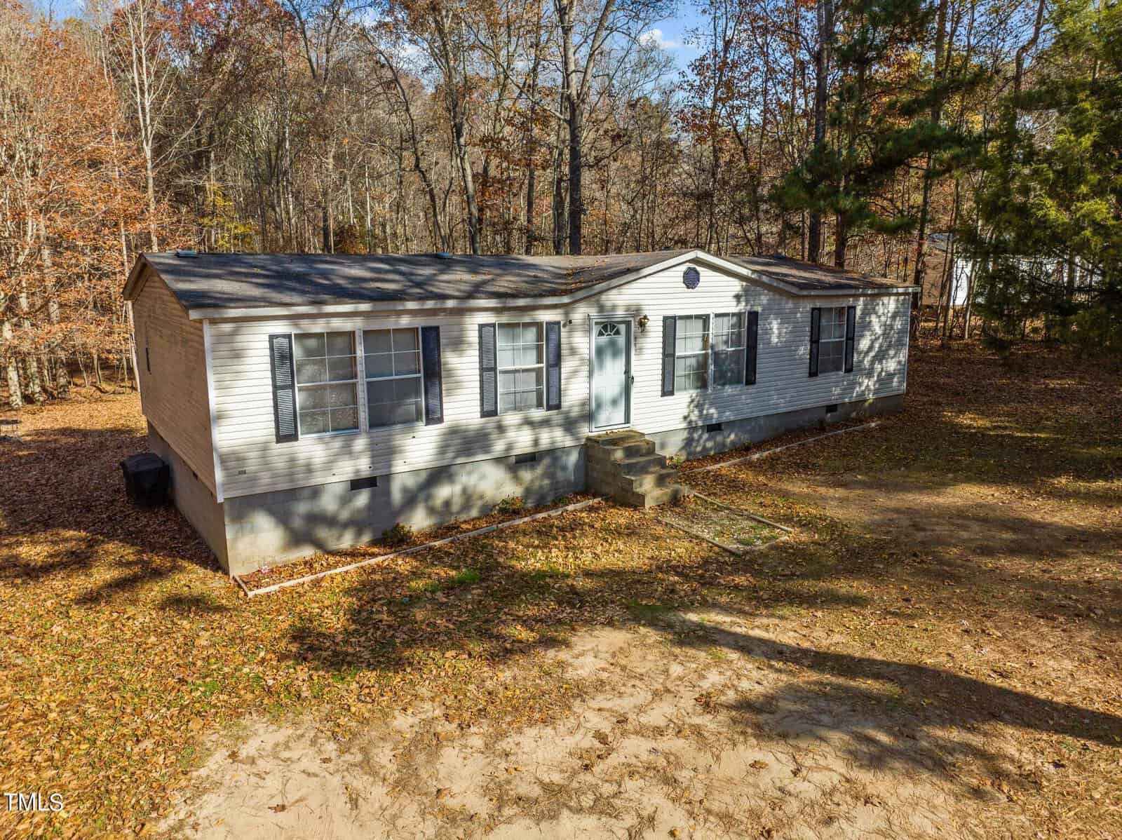 Aerial view of a single-story modular home surrounded by sparse trees and dry grass, with a clear blue sky above. the house features a white exterior and gray roof.