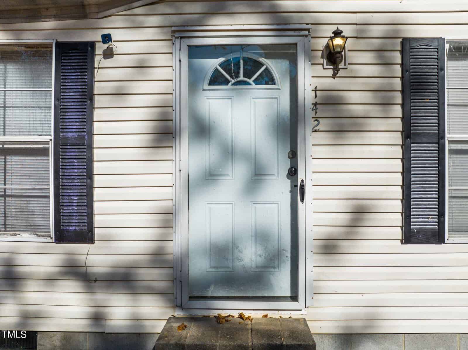 A front view of a residential building featuring a white front door flanked by two windows with black shutters, and a lantern-style light fixture on one side.