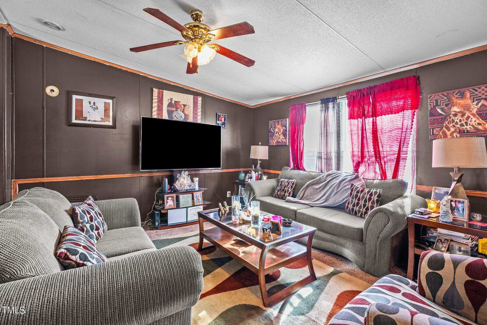 A cozy living room with dark brown walls and a patterned area rug. it features two sofas, a coffee table, a mounted tv, and a ceiling fan. bright red curtains adorn the window, and various decor items are visible.