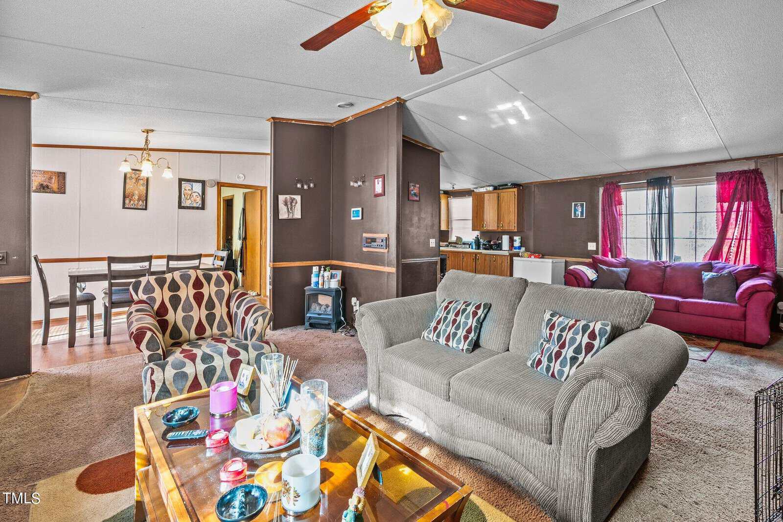 A spacious living room features a gray sofa and patterned armchairs, a glass coffee table, a dining area in the background, and an adjoining kitchen. bright decor with wood paneling and curtains.