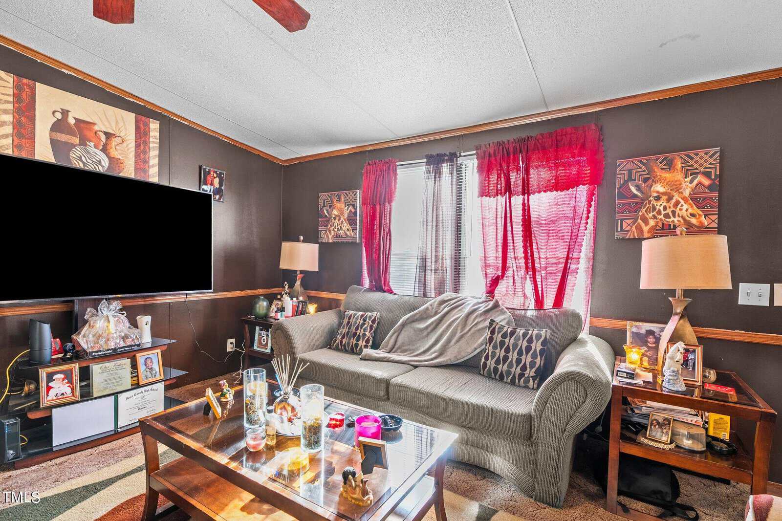 Cozy living room with a large sofa, red curtains, a glass coffee table, and cultural decorations on the walls. a big screen tv is mounted on one side.