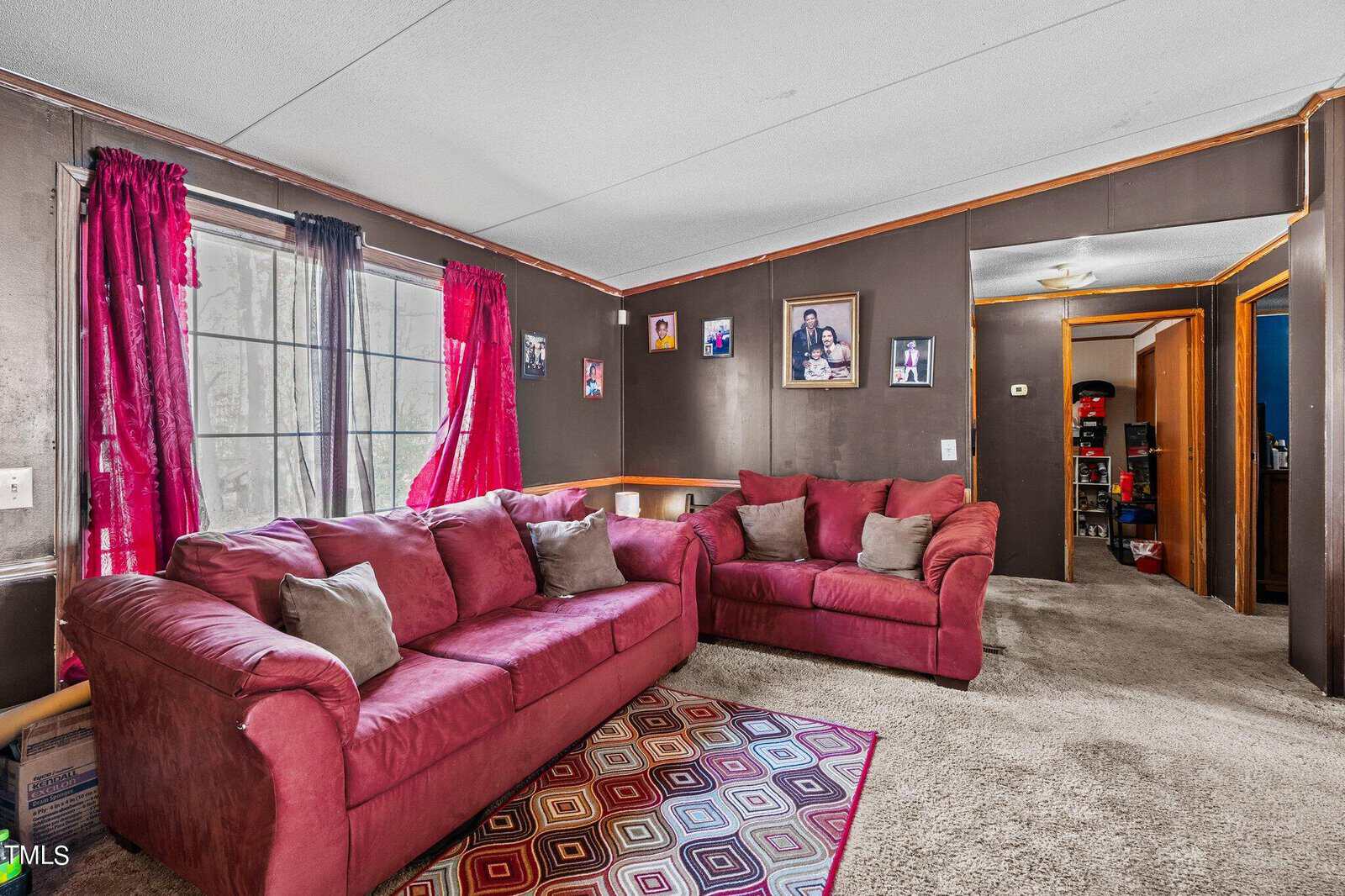 A cozy living room featuring a maroon l-shaped sofa with gray cushions, colorful rug, gray walls adorned with framed photos, and burgundy curtains beside an open door.