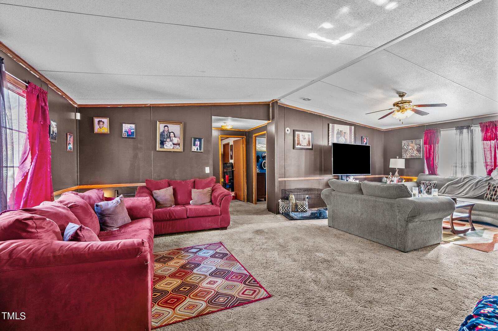 A spacious living room with a red sectional sofa, a gray couch, and colorful curtains. photos on the wall, carpeted floor, and a ceiling fan overhead. bright natural light illuminates the room.