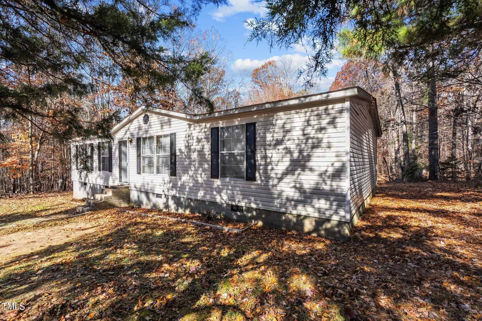 A single-story mobile home with beige siding, located in a wooded area with scattered fallen leaves and pine trees, under a clear blue sky.