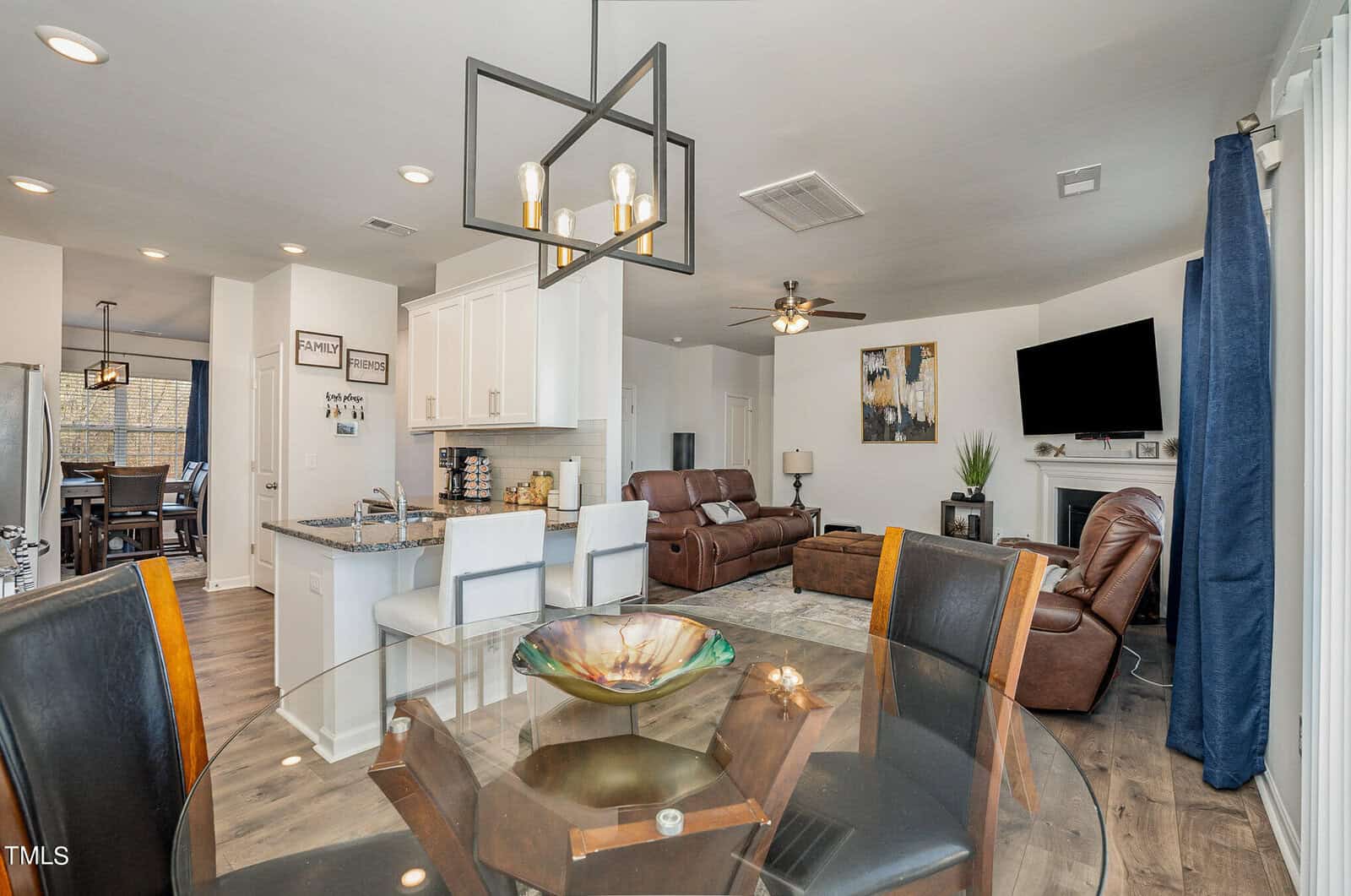 A modern open-concept living space with a dining area featuring a glass table and dark chairs, a kitchen with white cabinets and bar stools, and a living room with brown leather sofas and a wall-mounted TV above a fireplace. A stylish light fixture hangs above.