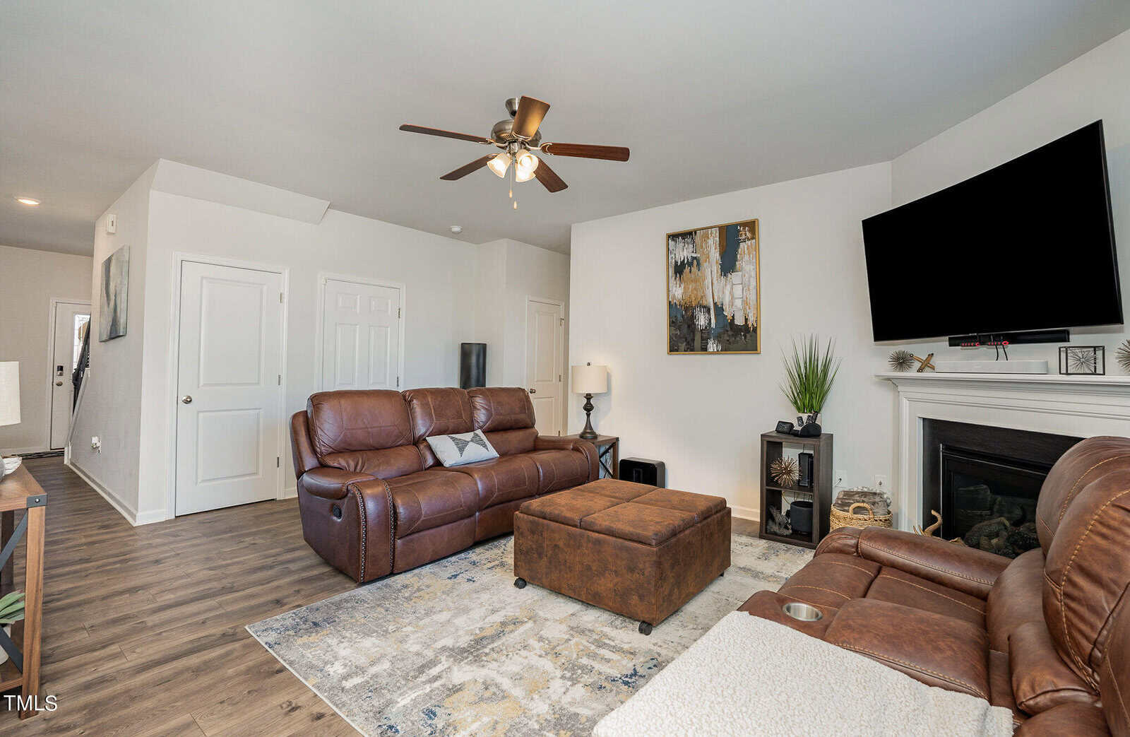 A cozy living room with brown leather sofas, a large ottoman, and a flat-screen TV mounted above a fireplace. The walls are adorned with modern artwork, and a ceiling fan hangs above. Wooden floors and natural light complete the inviting space.