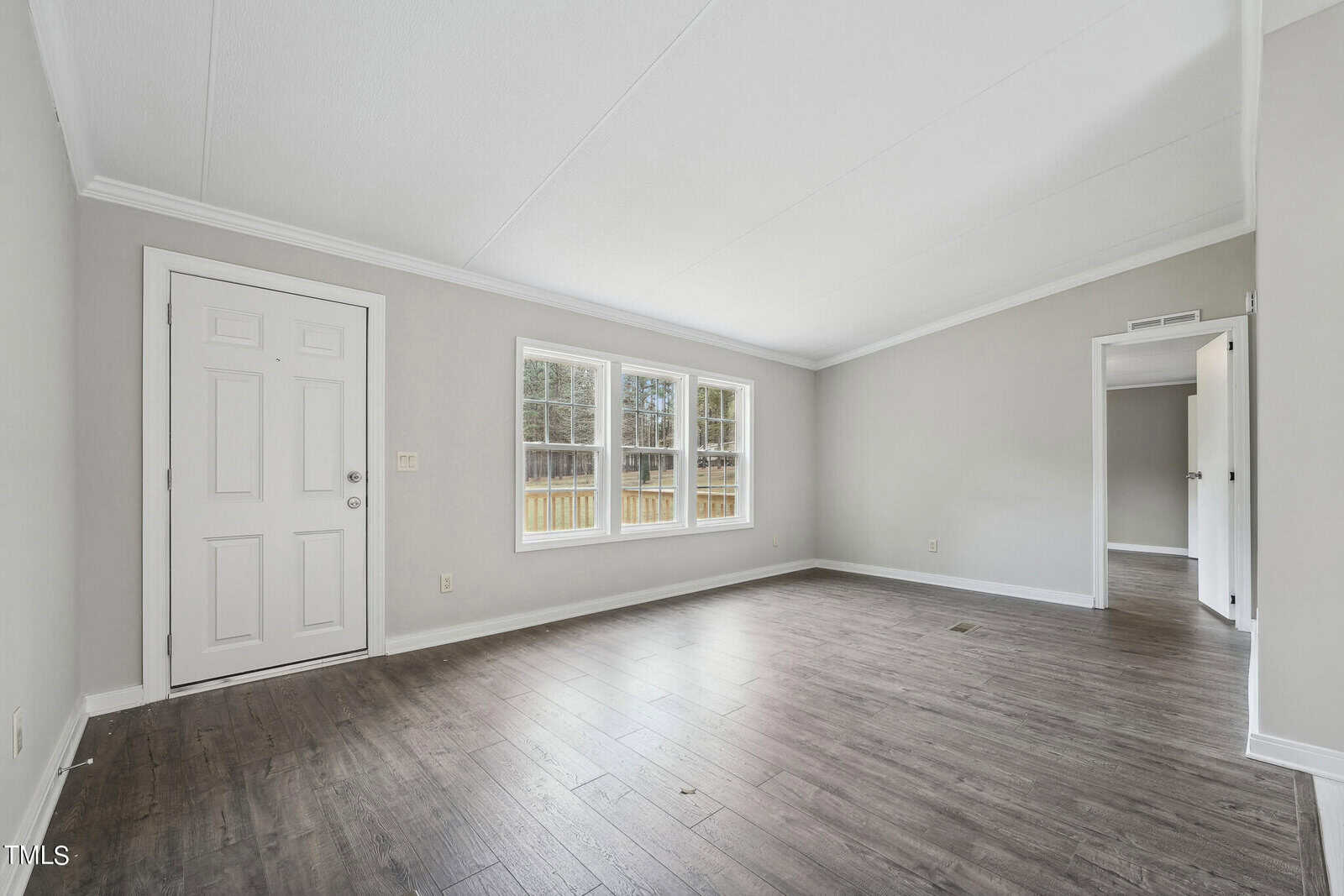 An empty room with hardwood flooring featuring a series of windows along one wall, a white entrance door on the adjacent wall, and an open doorway leading to another room.