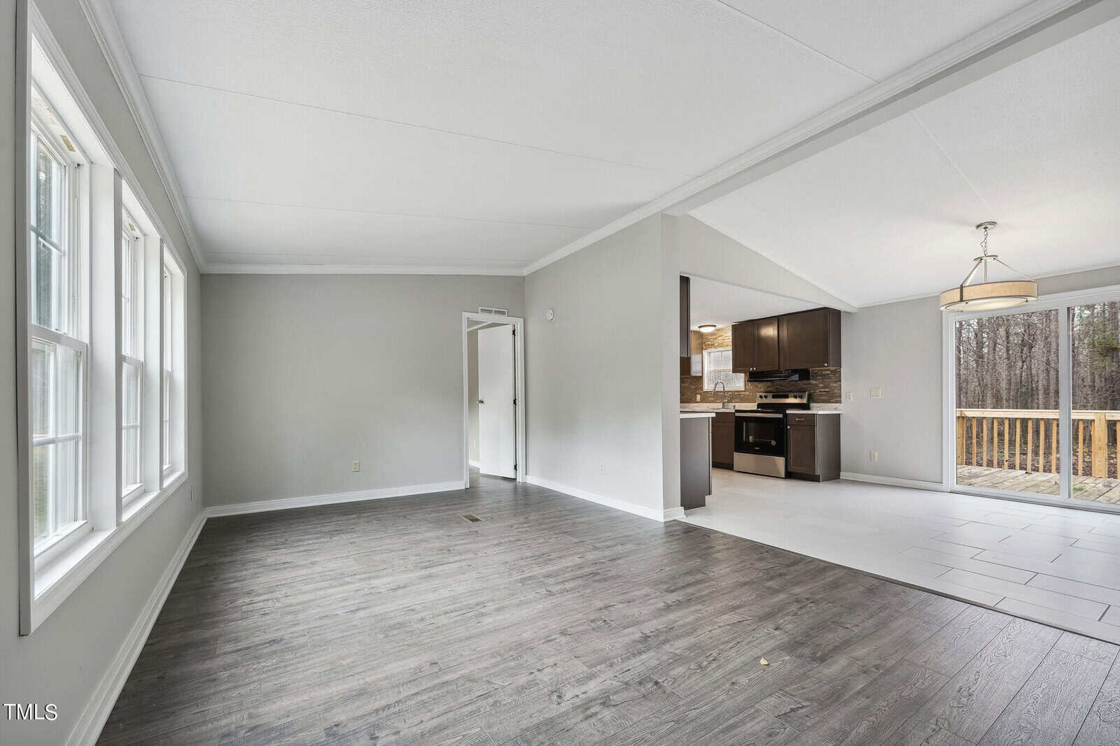 Spacious, unfurnished room with gray walls and dark wood flooring, flowing into a kitchen area with brown cabinets and stainless steel appliances, adjacent to a doorway leading to a deck.