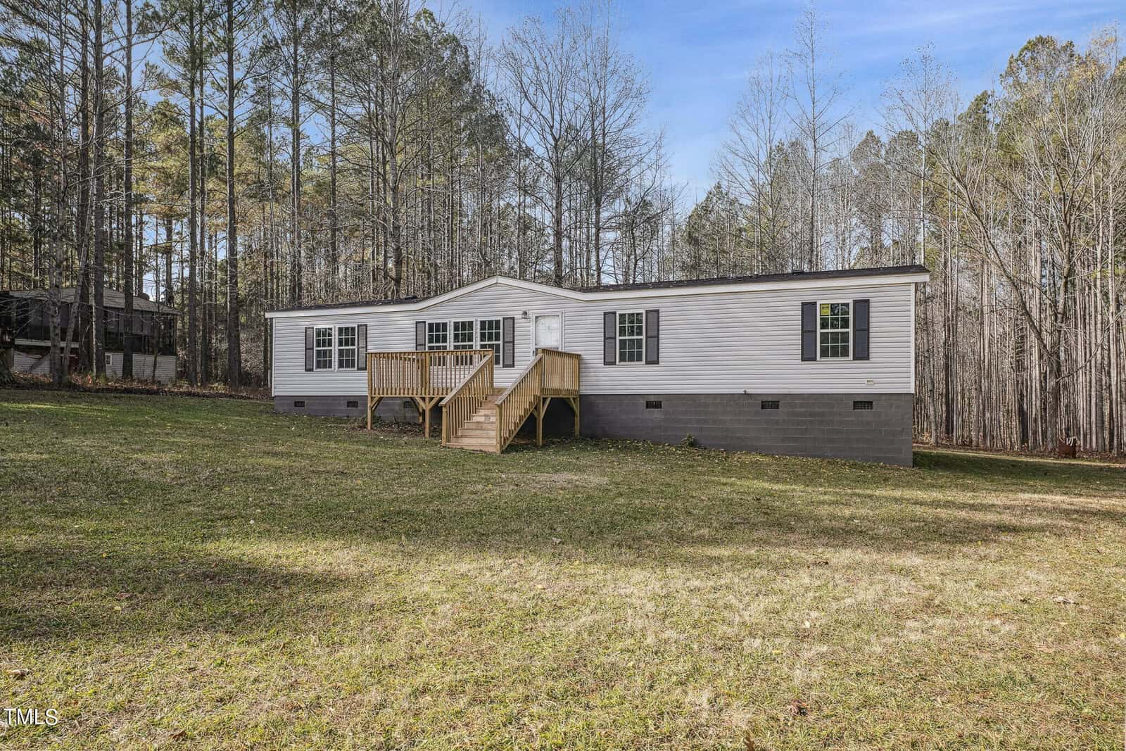 A gray single-story modular home with a small wooden deck and stairs, surrounded by a grassy yard and a backdrop of tall trees.