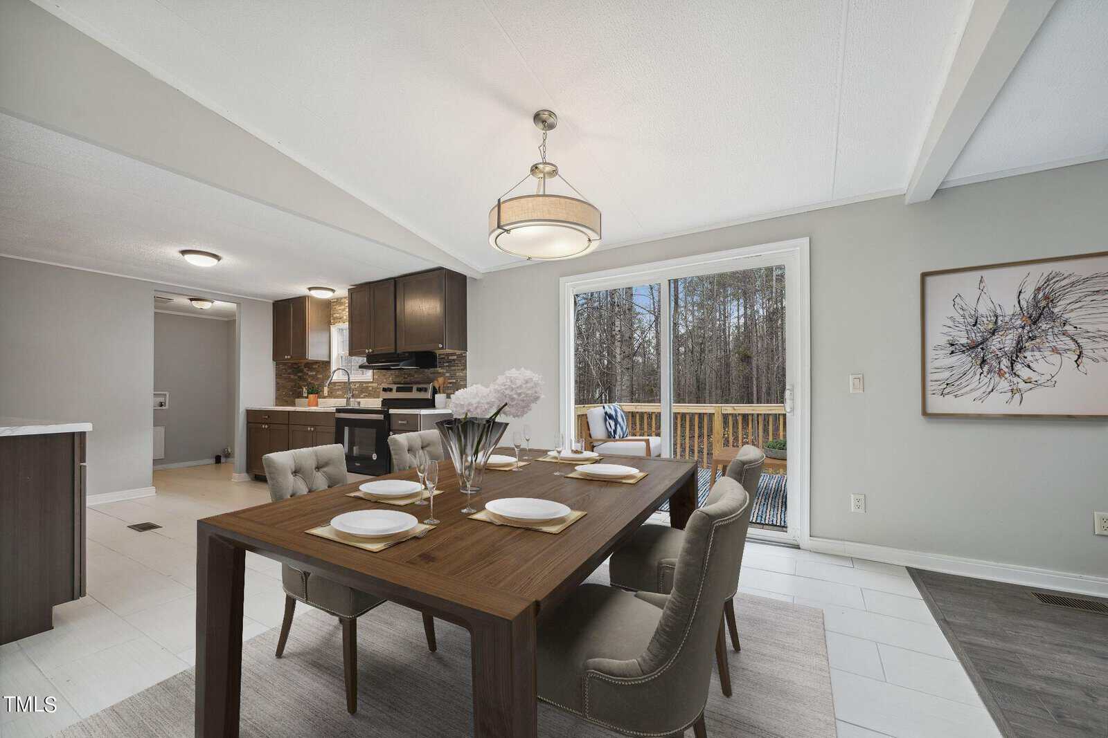 A modern dining area with a wooden table set for four, adjacent to an open kitchen with wooden cabinets, leading to a living space with a light-colored sofa set. the room has a large window providing a view of trees outside.