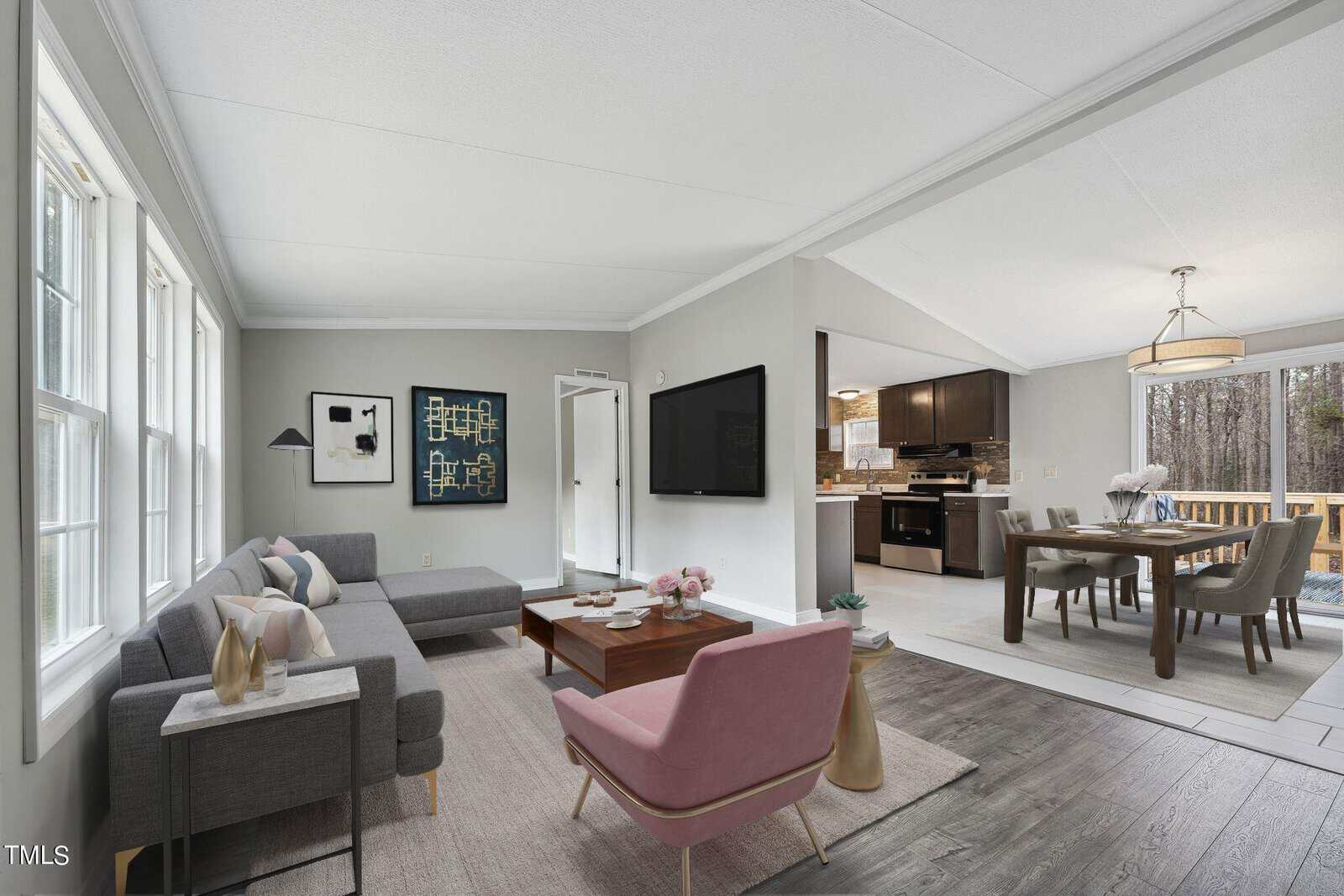 Modern open-plan living space featuring a seating area with a gray sofa and a pink armchair, a dining area with a wooden table and chairs, and a kitchen with dark cabinetry, connected by laminate flooring.