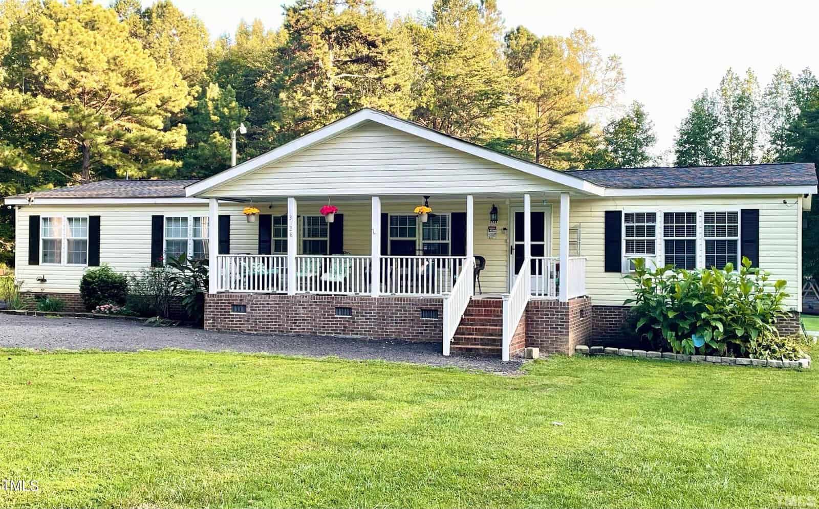 Located at 3128 Henry Wilson Road in Oxford, this single-story house boasts light yellow siding and a sturdy brick foundation. The covered porch is adorned with hanging plants, while the well-maintained yard features a lush green lawn and scenic trees as its backdrop.