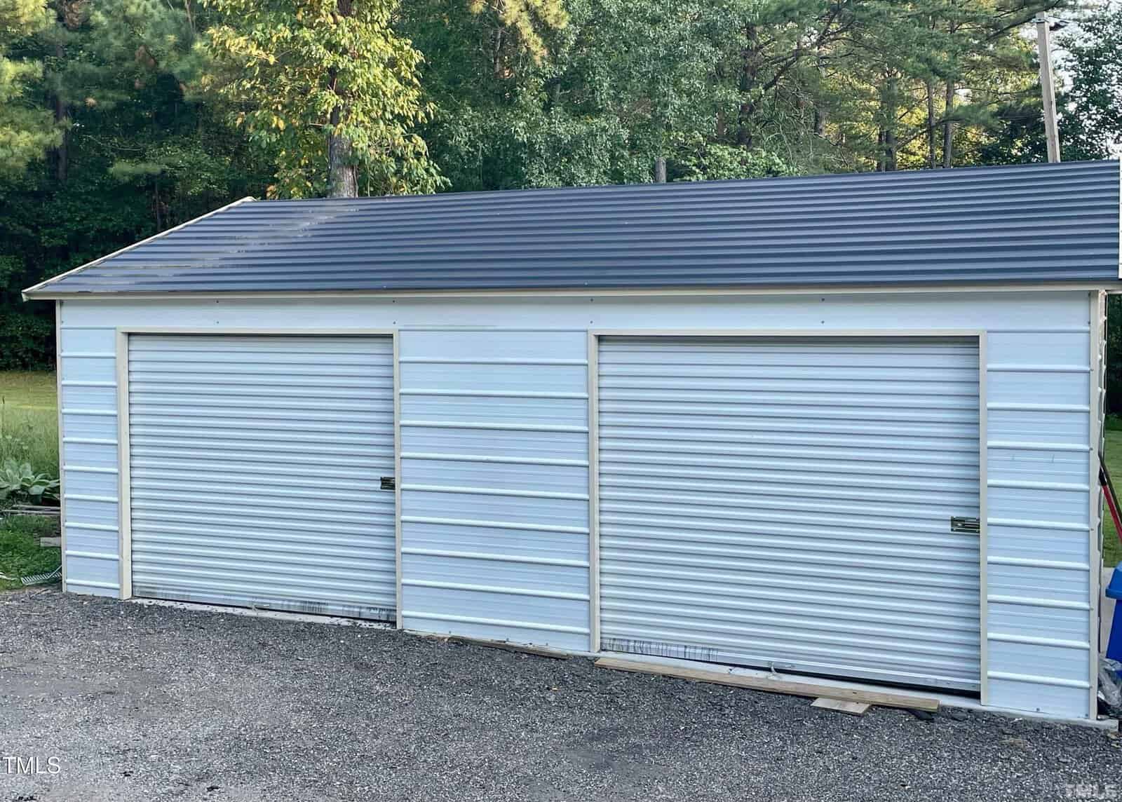 A white metal double garage with two closed roll-up doors is set in a wooded area. The roof is pitched and made of dark material, contrasting with the light-colored siding. The garage is surrounded by trees and greenery.