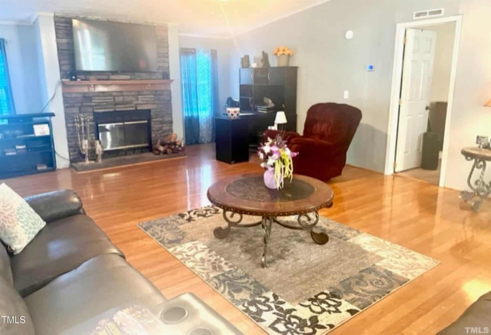 A cozy living room with a stone fireplace, a large TV mounted above, a dark leather sofa, and a plush red armchair. A round wooden coffee table with a flower vase sits on a patterned rug over polished wooden floors. A desk is in the corner near a door.