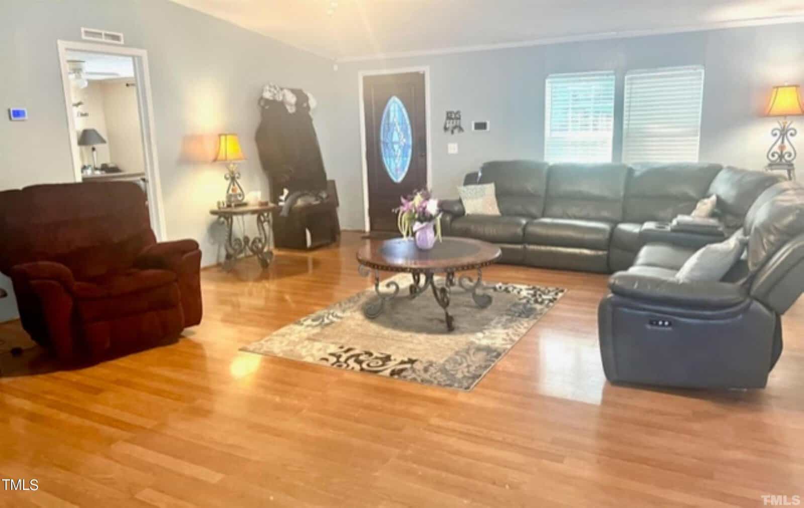 A cozy living room with a large sectional sofa, an armchair, a coffee table with flowers, and wooden flooring. Two lamps on side tables provide warm lighting. A decorative rug is beneath the furniture. A door with an oval glass window is visible.