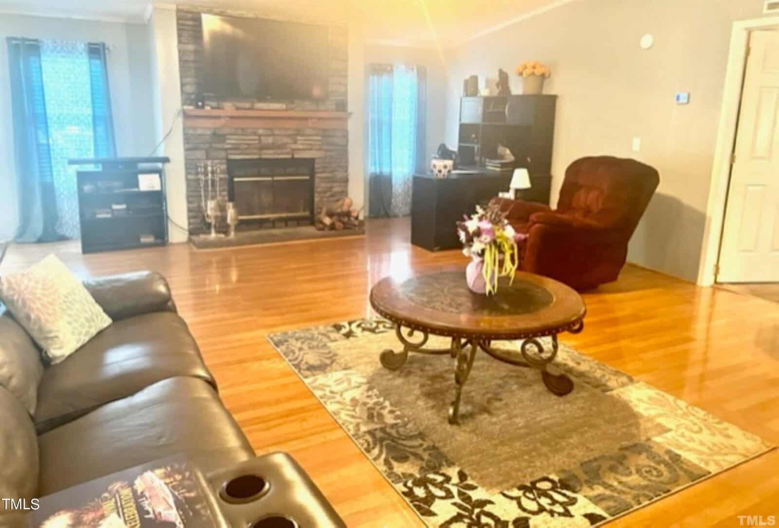 A cozy living room with wooden flooring features a brown leather couch, a round coffee table with flowers, and a large stone fireplace. A TV is mounted above the fireplace. A burgundy recliner and bookshelves are visible in the background.