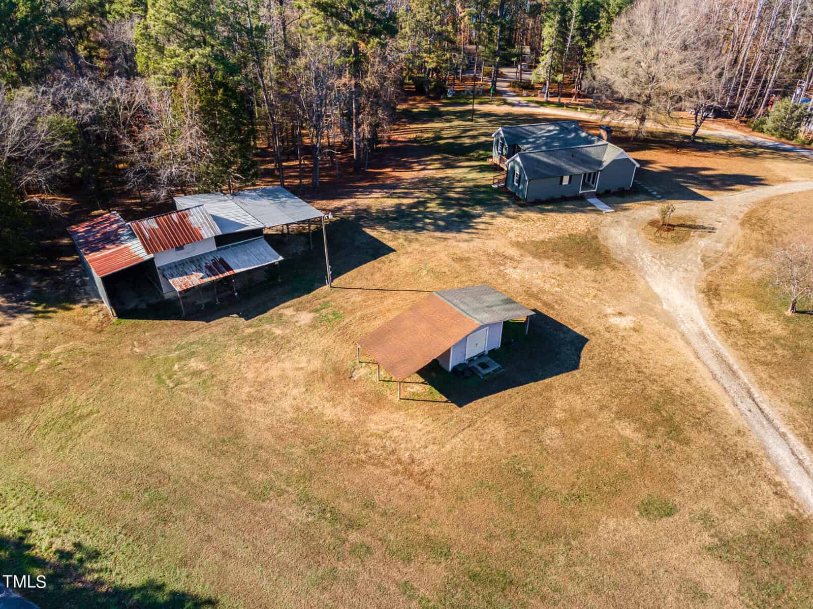 Aerial view of a rural property showing two buildings with rusty metal roofs and surrounding trees casting shadows, with a dirt path leading to the structures.