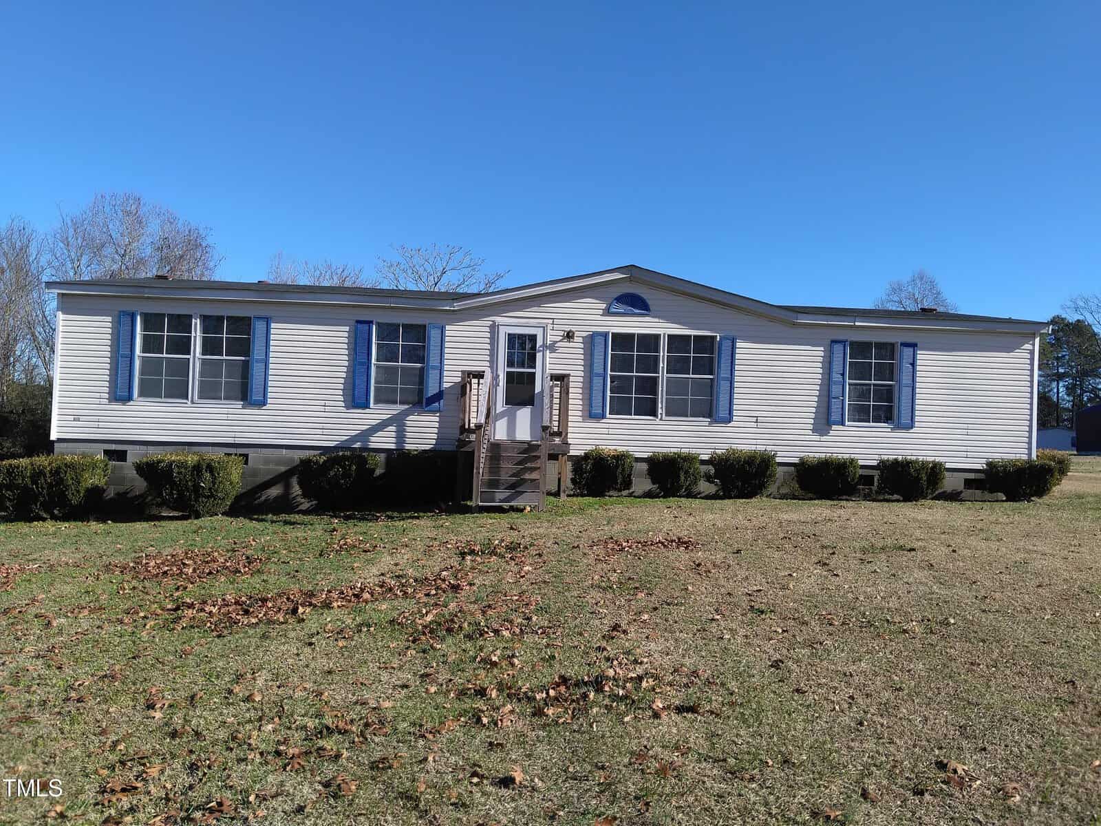 A single-story manufactured home with white siding and blue shutters, featuring central steps leading to the front door, situated on a grassy plot under a clear sky.
