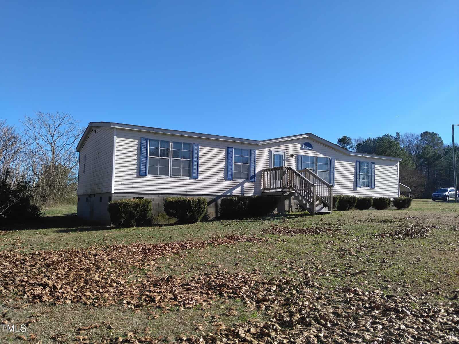 A single-story mobile home with a small wooden front porch situated on a grassy plot with scattered leaves under a clear blue sky.
