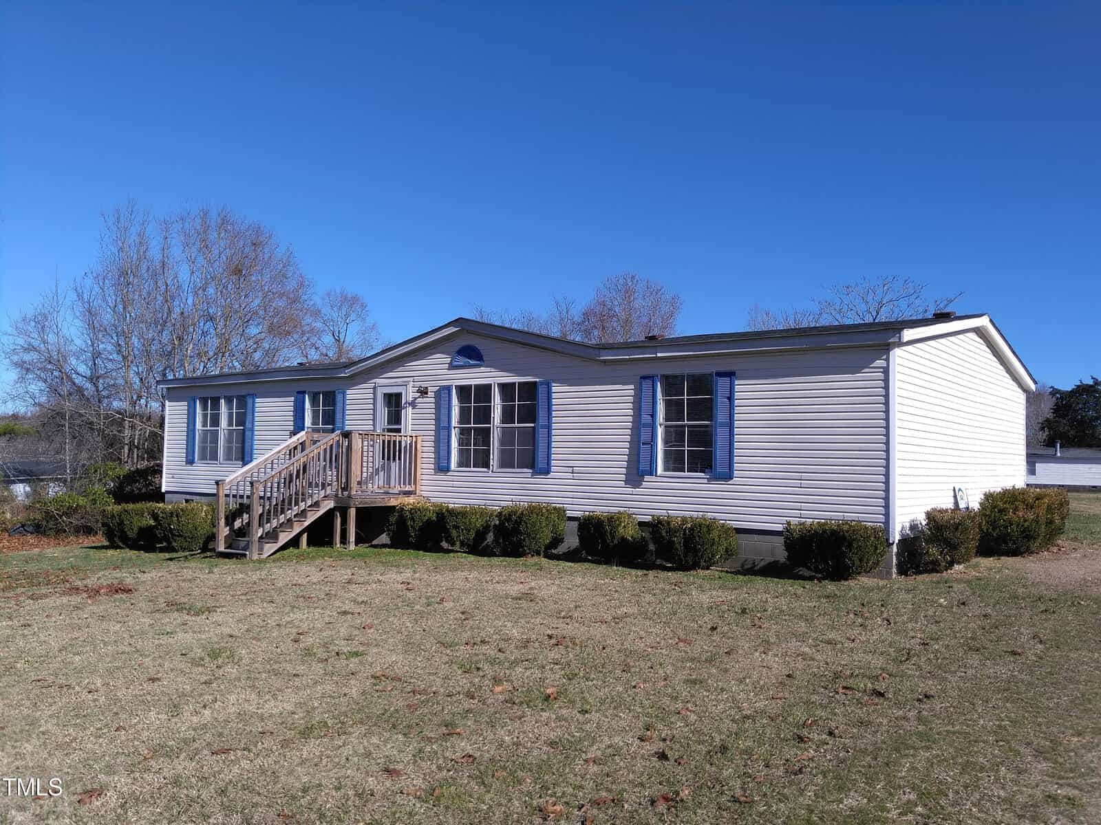 A single-story modular home with gray siding and white trim, featuring a small wooden front porch with steps, situated on a lawn with sparse grass under a clear blue sky.